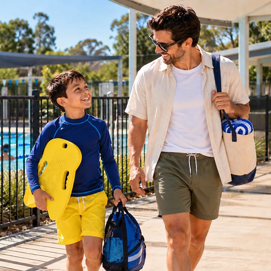 Parent and young boy arriving for an outdoor swimming lesson with a kickboard and swim bag.